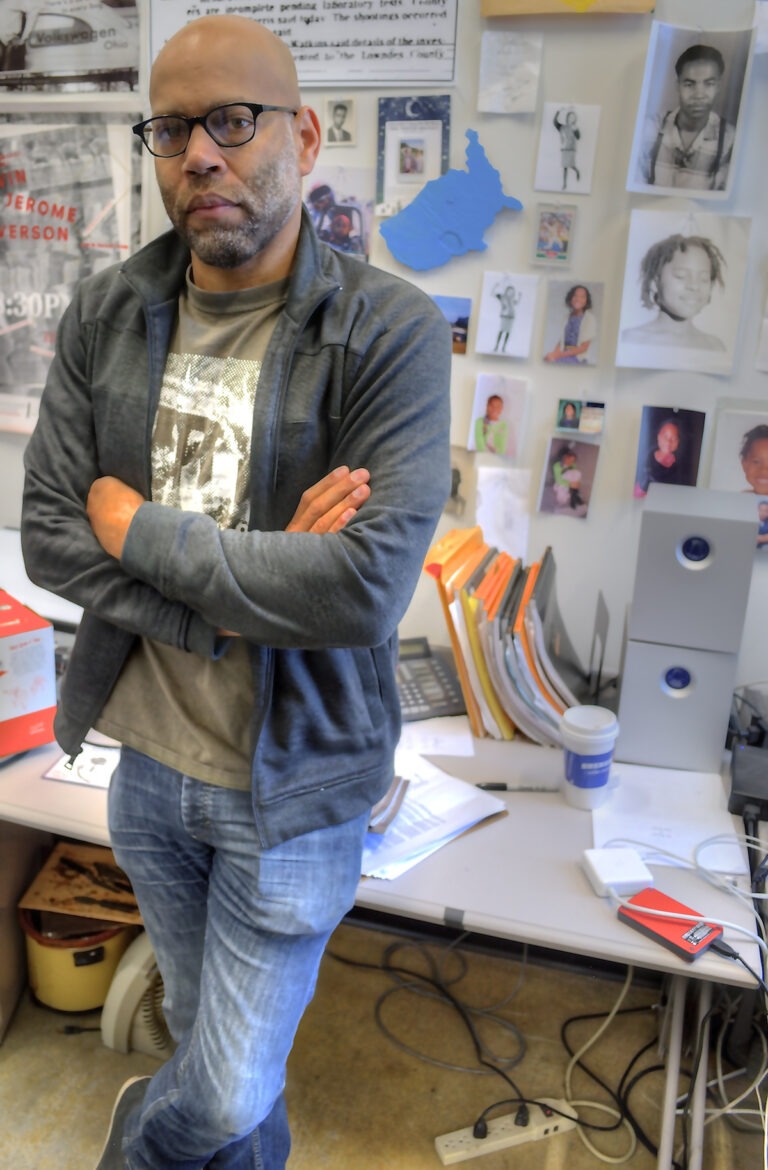Photograph of filmmaker Kevin Jerome Everson. He's a Black man, balding, wearing glasses and standing in his studio with his arms crossed