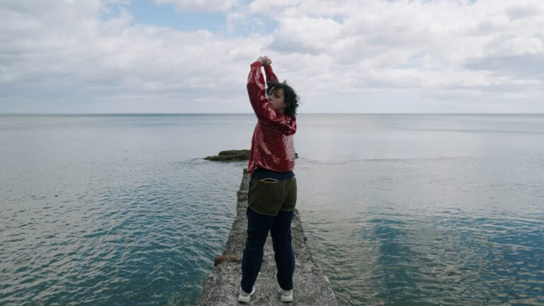 On a strip of concrete that juts out into the sea, a person wearing a red sparkly jacket stands tall with their arms stretched up and twists around to face the camera