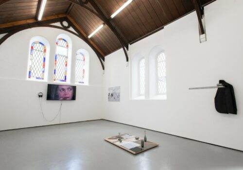 An art exhibition featuring a monitor showing a film of a woman's face, a painting, a wall installation made up of a jacket, and a floor sculpture that includes wood and glass. The gallery is a converted historical building with white walls, an arched wooden ceiling and stained glass windows.