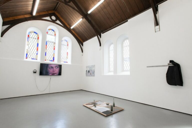 An art exhibition featuring a monitor showing a film of a woman's face, a painting, a wall installation made up of a jacket, and a floor sculpture that includes wood and glass. The gallery is a converted historical building with white walls, an arched wooden ceiling and stained glass windows.