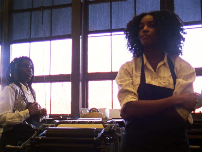 A still from the film ‘Pride’ by Kevin Jerome Everson and Claudrena N. Harold. The image features two Black young adults with long dark hair wearing darkly coloured aprons and white shirts with the sleeves rolled up. They are standing by a printing machine. The young adult closest to the camera is facing the camera with her arms folded to the right of the frame, looking to her right. The young adult to the left of the frame and in the background is side on to the camera and is looking straight ahead. In the background, there are large windows revealing sunlight, with blue blinds pulled down slightly and wooden framing.