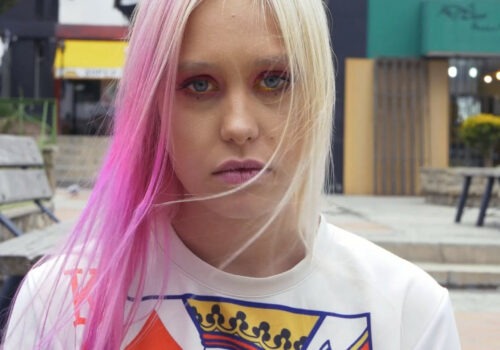 A person with light skin and long blonde straight hair dyed bright pink in sections wears matching pink and gold make-up