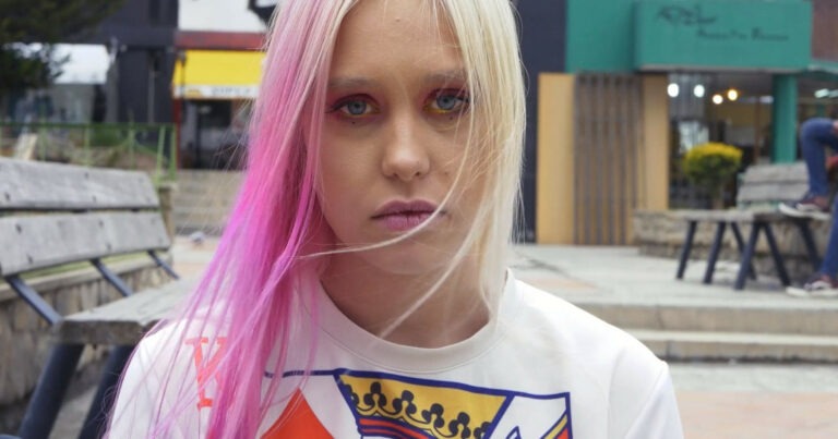 A person with light skin and long blonde straight hair dyed bright pink in sections wears matching pink and gold make-up