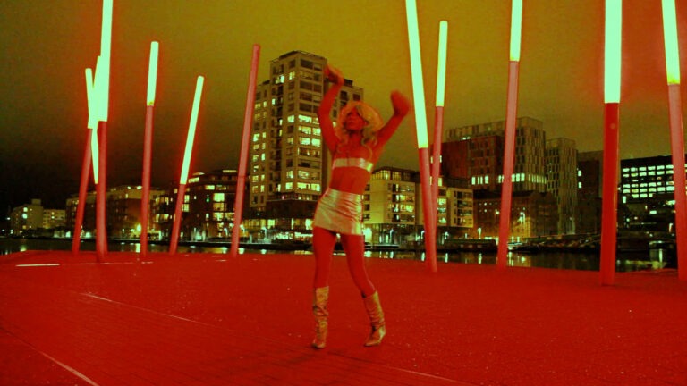 Venus Patel, wearing a gold two-piece mini skirt and belt top with gold boots, poses at the docklands in Dublin. The architectural lighting and city lights behind shine brightly the dramatic red-coloured ground that Venus is standing on reflects red colour onto her body