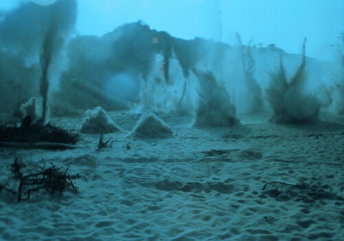 A still image from a Polish science fiction film by director Andrzej Żuławski, titled: On the Silver Globe, from 1988. The image shows a desolate landscape on an uninhabited planet, completely covered in a green-blue hue. In the foreground we see sand and branches on the ground. In the background, clumps of sand appear to burst up into the air from various positions. A mountainous or hilly landscape can be seen in the distance.