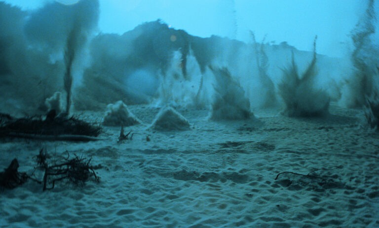 A still image from a Polish science fiction film by director Andrzej Żuławski, titled: On the Silver Globe, from 1988. The image shows a desolate landscape on an uninhabited planet, completely covered in a green-blue hue. In the foreground we see sand and branches on the ground. In the background, clumps of sand appear to burst up into the air from various positions. A mountainous or hilly landscape can be seen in the distance.