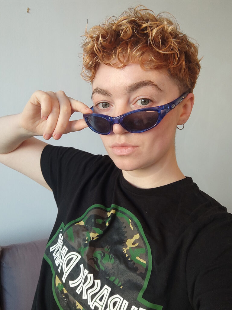 A colour headshot of artist Renèe Helèna Browne. Renèe is a white Irish person with short curly hair, they’re wearing a dark Jurassic Park T-shirt and are looking over their blue wrap-around glasses at the camera.