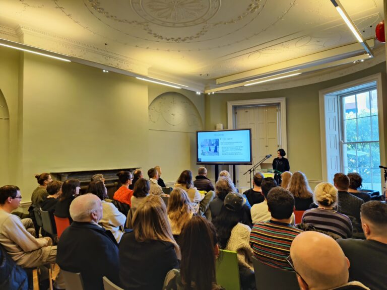 Fionnuala Sweeney, Head of Film and Architecture at the Arts Council of Ireland, is presenting an information session. She is standing in front of a microphone in the background of the image. There is a large modern television to her right, which displays her presentation. She has light skin, medium length dark hair, and wears black clothing. In the foreground there is a full room of audience members seated and observing her presentation, with their backs to the camera.