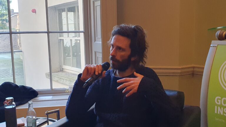 A photo of an Interview with film artist Tadhg O’Sullivan. He is seated and turned slightly away from the camera to his right. He is speaking into a microphone, which he holds in his right hand, and is gesturing with his left hand. He has light skin, dark hair, a dark beard, and wears a dark blue wollen jumper. He is cropped out of the image from the waist down. There is a cream wall behind him and a large window behind him to his right.