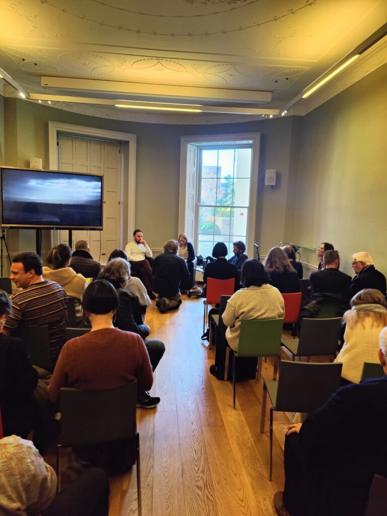 An Arts Council of Ireland Funding Clinic at Goethe Institute, Dublin, Ireland. A room full of people seated with their backs to the camera. To the left in the foreground there is a modern television standing. In the centre of the image in the foreground are two men and one woman who are leading the public discussion. There is a large window behind them.