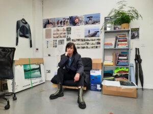 A studio portrait of filmmaker Lisa Freeman taken at Temple Bar Gallery + Studios in Dublin, Ireland. Lisa is seated on a cushioned chair in the center of the image. Behind her, various images from her film work and other photographs are visible. To Lisa's left, there is a shelf with many notebooks and a plant. Lisa is wearing green and black boots, dark trousers and a dark blazer, with a blue tshirt. Her posture is relaxed but confident, as she leans forward with her legs apart and rests her chin on her right hand while looking directly into the camera.