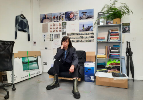A studio portrait of filmmaker Lisa Freeman taken at Temple Bar Gallery + Studios in Dublin, Ireland. Lisa is seated on a cushioned chair in the center of the image. Behind her, various images from her film work and other photographs are visible. To Lisa's left, there is a shelf with many notebooks and a plant. Lisa is wearing green and black boots, dark trousers and a dark blazer, with a blue tshirt. Her posture is relaxed but confident, as she leans forward with her legs apart and rests her chin on her right hand while looking directly into the camera.