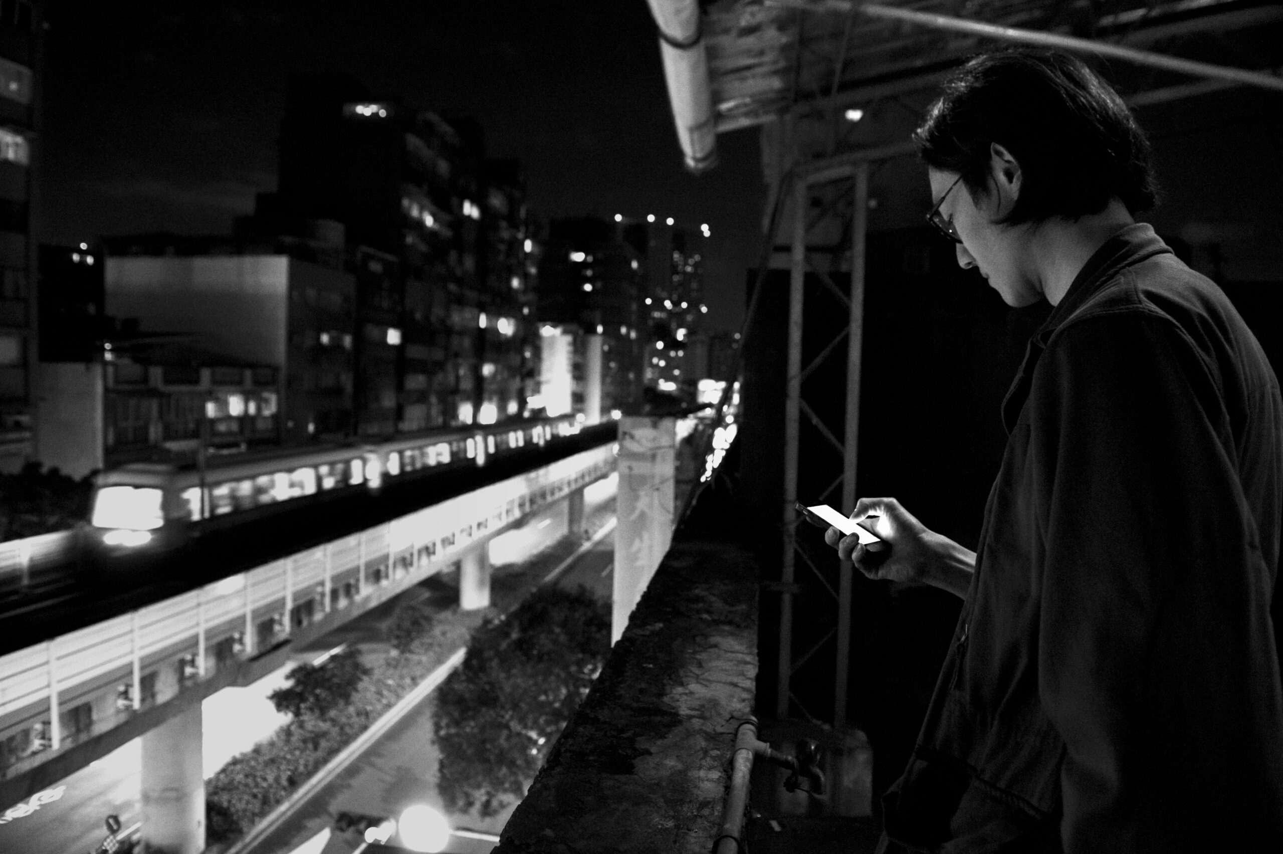 A black and white still from the film 'Far Away Eyes' by the director Wang Chung-Hong. In this image, we see a Taiwanese man in his late 20's standing on a balcony of a building located in an industrial area in Taiwan at night-time. He is wearing glasses and a dark jacket, and he is standing to the right of the frame, slightly turned away from the camera. In his right hand, he holds his mobile phone, which he is focusing on. The man is facing railway tracks, and we can see a train passing by in the background. Behind him, there are several apartment blocks and some shrubbery.