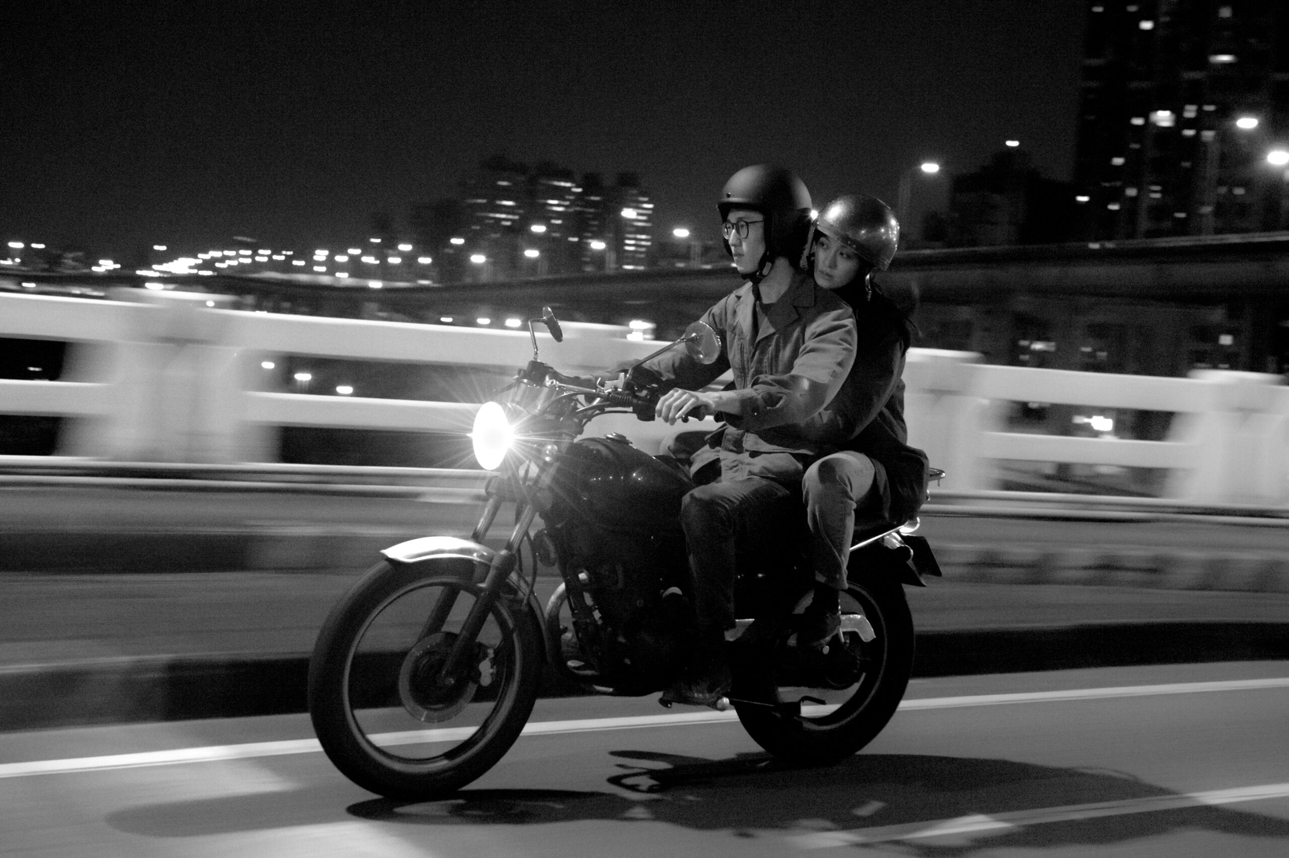 A black and white still from the film 'Far Away Eyes' by the director Wang Chung-Hong. In this image, we see a Taiwanese man in his late 20's driving a motorcycle at night-time, with a young woman of a similar age as his passenger holding onto him from behind. The man wears glasses, and they are both wearing dark jackets, trousers, and helmets. The cityscape in the background is blurry, creating a sense of motion.