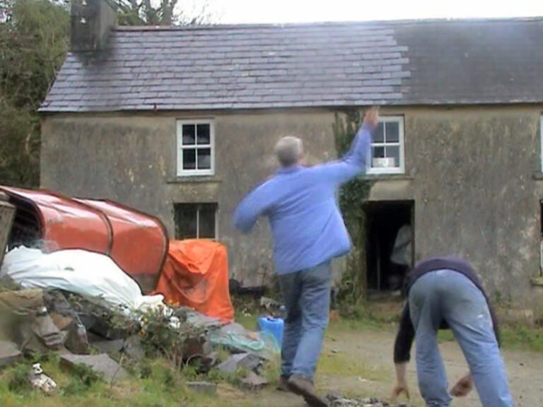 A still from the short film Windows Blowing Out (2005) by Jorge Satorre. The image depicts two men standing in front of a country house in a rural area. The man in the centre of the image has short grey hair and is wearing blue jeans and a blue shirt. His right arm is raised, appearing to throw something, and he is holding a stone in his hand. The man to the right of the frame is bending forward and picking up stones. He is wearing blue jeans and a long-sleeved dark top. In the background, there are larger rocks and orange-coloured materials. The house is grey with a dark tiled roof, and the area in front of the house where they are standing is grassy.