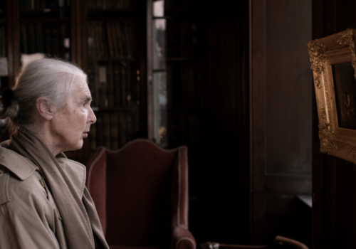This image is a still from the short film 'A Moment Twice Lived' by Irish filmmaker Martin Healy. The image depicts an elderly woman seated with her side to the camera, gazing at a painting. She wears a beige mack jacket, a beige scarf, and has her grey hair tied in a bun. In the background, there is a red antique chair wooden bookcase and wooden window shutters. The painting has a golden, antique-style frame with flowers, and appears to show the silhouette of a person standing against a fiery background, although it is unclear due to the angle. The woman appears to be deeply absorbed in the painting.