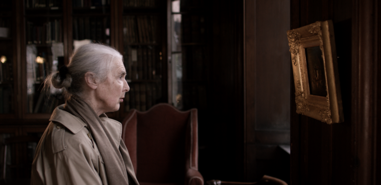 This image is a still from the short film 'A Moment Twice Lived' by Irish filmmaker Martin Healy. The image depicts an elderly woman seated with her side to the camera, gazing at a painting. She wears a beige mack jacket, a beige scarf, and has her grey hair tied in a bun. In the background, there is a red antique chair wooden bookcase and wooden window shutters. The painting has a golden, antique-style frame with flowers, and appears to show the silhouette of a person standing against a fiery background, although it is unclear due to the angle. The woman appears to be deeply absorbed in the painting.