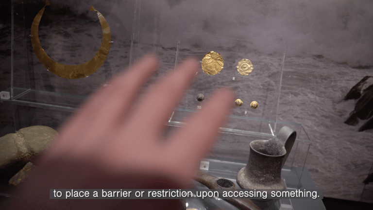 This still image is from the 2021 film ‘No One Can Ever Embargo the Sun’ by artist and filmmaker Amanda Rice. The image portrays a blurry hand reaching towards two clear panels, which encase gold crescent and circular shapes of various sizes that are reminiscent of ancient jewellery and artefacts. In the mid-ground, there are several objects including a jug and a rock, both with a similar archaic style. The background features a grey, dusty landscape. At the bottom of the screen, there is white text displayed on a grey border that reads 
