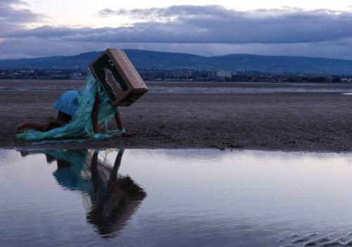 This is a still from ‘Daisy: Queer Prophet of the Apocalypse’, a work-in-progress film by artist and filmmaker Venus Patel. The scene is set at dusk on a sandy beach, where a person is shown on their hands and knees, facing sideways towards a low pool of water. They are dressed in a bright blue skirt, and on their head, there is a large rectangular cardboard box painted with a crocodile head. The box is adorned with long green and blue streamers, and their image is reflected in the still water. In the background, we see a townscape and low hills under a cloudy grey sky, with the sun breaking through the clouds in the distance.