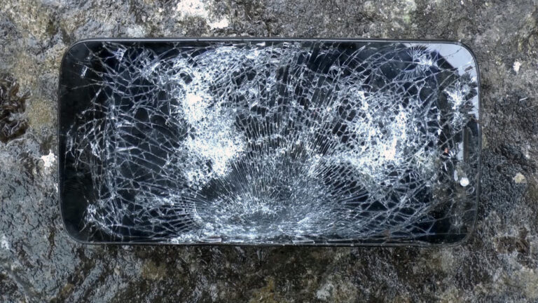 A still from the short film What Is An Apparatus? (2016) by Sean Lynch. The image depicts a black mobile phone lying on a concrete ground. The phone's screen is completely shattered.