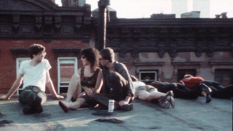 A still image from the film "New York Our Time" (2020) directed by Vivienne Dick. The scene features a group of adults sitting on a rooftop. Three of them are lying on their stomachs, soaking up the sun, while the other three are having a casual conversation. One man can be seen holding a can of Budweiser. The overall tone of the image has a vintage aesthetic.