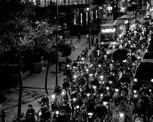 A black and white still from the film 'Far Away Eyes' by the director Wang Chung-Hong. In this image, we see a night-time street filled with motorcycles, along with a few cars and a bus trapped in traffic. To the left of the frame, we can see some houses and a few people on the pavement. Trees are planted along the sidewalk. The scene has a slightly ominous atmosphere.