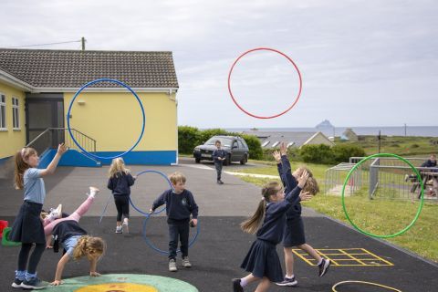 A still from the feature film 'Fields of Darkness' by Irish Michael John Whelan. The image depicts a school playground in rural Ireland. In the forefront of the image we can see a group of young primary school children playing together with brightly coloured hula hoops. They are wearing dark coloured jumpers, and trousers or skirts. In the background we can see a section of the school which is painted yellow and blue, as well as fields, houses, and the coastline.