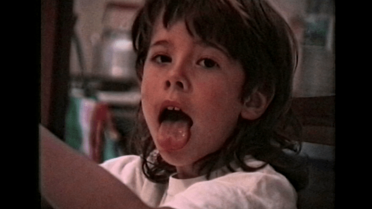 This is a still from the short film 'Yon' (2021) by Bárbara Lago. The image depicts the face, neck, and shoulders of a young child with a mullet haircut, brown hair, olive-skinned, and brown eyes. The child is looking directly at the camera with their tongue sticking out, and their two front teeth are visible, with a small gap between them. Their body is turned slightly towards their right side, and their left arm is partially visible in the bottom left of the frame. In the background, we can discern some household items, although they appear blurry. This still was captured on VHS.