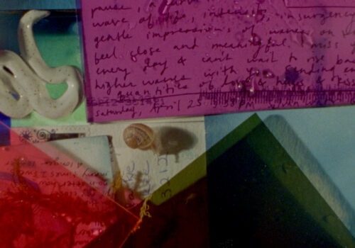 This is a still from Sofia Theodore-Pierce's short film 'Other Tidal Effects' (2021), featuring handwritten materials on overlapping cyanotype treated postcards. In the top half, a pink transparent plastic sheet with water droplets is placed over spidery handwriting. To the top left of the frame, there is a ceramic snake placed on light green and blue paper. On the bottom left-hand side of the frame, there is a transparent red plastic sheet with water droplets. It sits on small pieces of dried seaweed, as well as partially visible handwriting and a pale yellow tinted postcard with a stamp. A snail poking its head out of its shell is also visible crawling on top of the postcard, left of centre. On the bottom right-hand side of the frame, there is a dark green transparent plastic sheet that partially overlaps some of the materials on the bottom left. Beneath everything, there is a mottled blue sheet of paper, which is visible in the top left corner and bottom right of the frame.