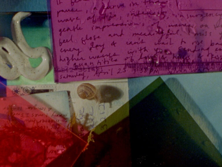 This is a still from Sofia Theodore-Pierce's short film 'Other Tidal Effects' (2021), featuring handwritten materials on overlapping cyanotype treated postcards. In the top half, a pink transparent plastic sheet with water droplets is placed over spidery handwriting. To the top left of the frame, there is a ceramic snake placed on light green and blue paper. On the bottom left-hand side of the frame, there is a transparent red plastic sheet with water droplets. It sits on small pieces of dried seaweed, as well as partially visible handwriting and a pale yellow tinted postcard with a stamp. A snail poking its head out of its shell is also visible crawling on top of the postcard, left of centre. On the bottom right-hand side of the frame, there is a dark green transparent plastic sheet that partially overlaps some of the materials on the bottom left. Beneath everything, there is a mottled blue sheet of paper, which is visible in the top left corner and bottom right of the frame.
