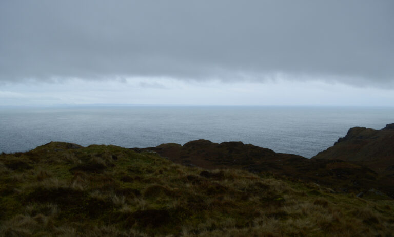 This is a still from the film 'Eyes Like Cats' (2022) by artist and filmmaker Susan Hughes. The scene depicts a rugged Irish landscape, with a cliff edge overlooking the sea under an overcast sky. In the foreground, there is an area of dark green grass, and rocky cliffs covered in dark green and brown moss. At the centre of the image, a grey-blue sea is visible, with light ripples on its surface. In the background, there is a light grey-blue horizon and grey clouds.