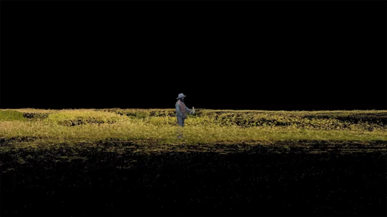 This is an image from the short film 'Constant' (2022) by Sasha Litvintseva and Beny Wagner. The image appears to be computer-generated or have a similar effect. The image depicts a man with dark skin and a dark-coloured beard standing in a field. He is holding what appears to be a piece of corn in his right hand and wearing a light blue sun hat, along with a light blue and brown full-length shirt and trousers. The corn field surrounds him and is coloured yellow and green. The sky in the background behind him and the foreground at the bottom of the image are completely black.