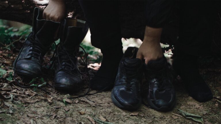 This is a still from Dan Guthrie's short film 'black strangers'(2022). The image depicts a man seated facing the camera on a low wooden branch in a forested area with dry leaves, foliage, and debris scattered on the muddy ground. In the frame, only his hands, feet, and lower legs are visible as he reaches forward, holding onto two pairs of shoes positioned on the ground. He is wearing a long-sleeved black top, black socks, and dark trousers. The shoes he is holding consist of men's lace-up black boots on the left side of the frame and black trainers on the right side. We can see sunlight filtering through the background behind him.