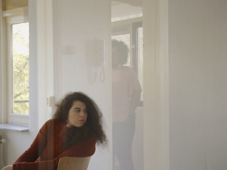 This still is from the film 'Two Stones' (2019) by Wendelien Van Oldenborgh. The image shows a woman seated in a white room on a wooden chair positioned to the left side of the frame. She is facing forward but has turned her head sideways towards the centre of the image. The woman has pale skin, long dark curly hair, and is wearing a burgundy-coloured jumper. To her right, there is a window through which trees are visible. The image gives the impression of being viewed through a large window, with a white door frame reflecting in the centre. In the door frame stands another woman with dark skin, shoulder-length brown curly hair, glasses, and wearing a pink t-shirt and jeans. Her hands are in her pockets. Behind her, there is another window with trees. The right side of the frame consists of a plain white wall.