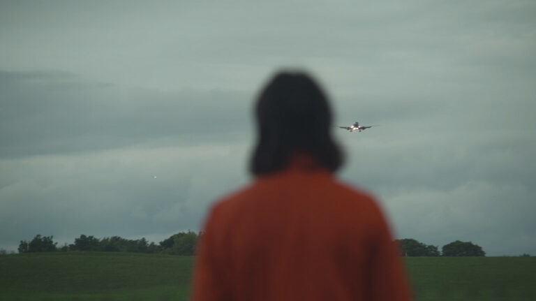 This is a still from the film 'Pet World' (2022) by Sofia Theodore-Pierce and Grace Mitchell. The image depicts a person standing in the foreground, viewed from behind, with shoulder-length dark hair and wearing a long-sleeved orange top. The person is slightly out of focus, and only the back of their head and upper back is visible within the frame. They appear to be looking at a low-flying plane preparing to land in the background. The sky has a somewhat dark and cloudy grey tone, and the plane's lights are turned on. The background consists of a dark green field in the lower part of the frame, with low bushes and trees at the edge of the grassy area. The entire background, including the field and trees, is in focus.