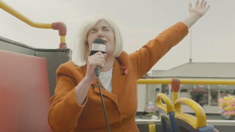 This is a still from the film 'Where's Danny' (2022) by Amy Pennington. The image depicts a person with light skin and a white-blonde bobbed wig. They are seated in an open-top bus, facing the camera. The person is dressed in an orange suit jacket with a white T-shirt underneath. Their left arm is extended diagonally in a gesture of exclamation, while their right hand holds a microphone up to their mouth. Their eyes are closed. In the background, there is a blurry shopfront with a collection of balloons outside. The interior of the bus is red, grey, and yellow in colour. The sky in the background is light grey.
