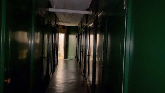 This is a still from the film 'Vagary' (2023) by Ross Hewitt. It portrays a dimly lit corridor resembling a school corridor, with an open door at the end that allows light to illuminate parts of the space. The light reflects off the darkly coloured concrete floor and on the rows of dark green lockers, which appear on either side of the frame. The ceiling between the lockers is white, and a lamp hangs from above but remains unlit. The image is captured at a slight diagonal angle.
