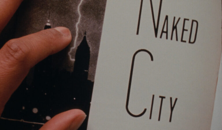 ​​This is a still from the film 'A Human Certainty' (2021) by Morgan Quaintance. The image shows the cover of the book 'Naked City' (1945) by photographer and photojournalist Weegee. On the right-hand side of the frame, we can see the words 'Naked City' in large black text on a white background. On the left-hand side of the frame, there is a black and white photograph of lightning hitting a tower on a New York City skyline at night. A hand can be seen partially on the left side of the frame, with the thumb, index finger, and part of the middle finger visible and touching the photograph.