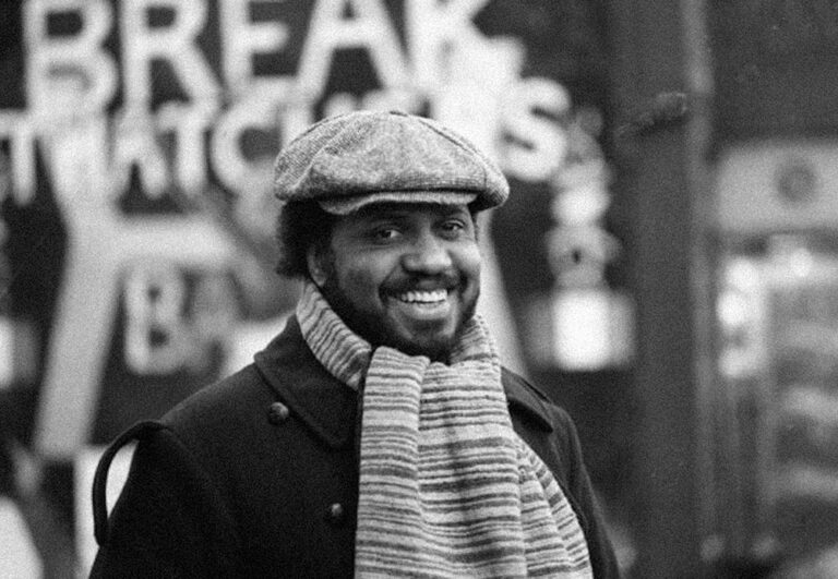This is a still image from the documentary film 'The Black and the Green' (1983) directed by St. Clair Bourne. The image features a black man with short dark hair and a short beard. He is facing the camera and smiling warmly, with only his head and upper torso captured within the frame. He is dressed in a dark-coloured winter coat, a light-coloured scarf with dark, thin stripes, and a light-coloured flat cap. The background appears blurred, and some large writing can be discerned. The entire image is presented in black and white.