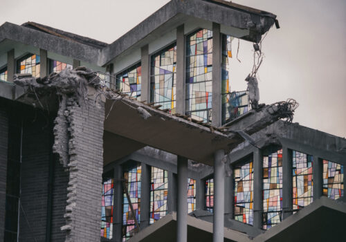 This is a still from the film ‘Making Dust’ by Irish filmmaker Fiona Hallinan. The image depicts a portion of the demolished ‘Church of the Annunciation’ in Finglas, Co. Dublin. A metal frame and fragments of grey brick wall support the remaining structure. The centre of the image features large stained glass windows with metal dividers. The stained glass is brightly coloured and comprised of small abstract panels in shades of blue, pink, yellow, green, red, and brown. The sky in the background is a light grey colour.