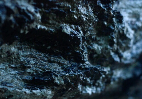 This image is a still from the work-in-progress film 'May the Hills and the Heights Have You' by Susan Hughes. It features a close-up shot of a wet rocky area with daylight highlighting sections of the rock. The rock is coloured in various tones of grey and black, and the right section of the frame is out of focus.