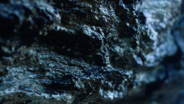 This image is a still from the work-in-progress film 'May the Hills and the Heights Have You' by Susan Hughes. It features a close-up shot of a wet rocky area with daylight highlighting sections of the rock. The rock is coloured in various tones of grey and black, and the right section of the frame is out of focus.