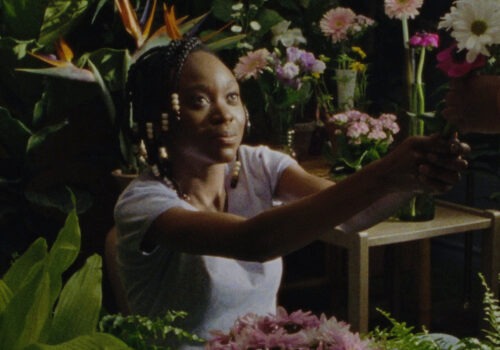 This is a still from the film 'Cette maison (This House)' by Haitian-Canadian director Miryam Charles. In the image, a person with dark skin tone is seated in a room filled with green plants and colourful flowers in shades of pink, purple, orange, and white. Their hair is braided and secured at the ends with cream and brown beads. They are dressed in a pale purple t-shirt and seated at a diagonal angle to the camera. Their gaze is directed at a person just outside the frame, with only their left hand visible. With both hands outstretched, they are handing a bunch of white flowers to the unseen individual.