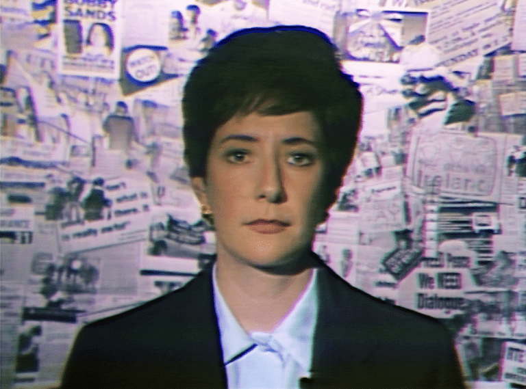 A woman with short black hair stands looking at the camera in front of a wall. The wall features a collage made from newspaper clippings relating to censorship. She is wearing an 1980s style suit.