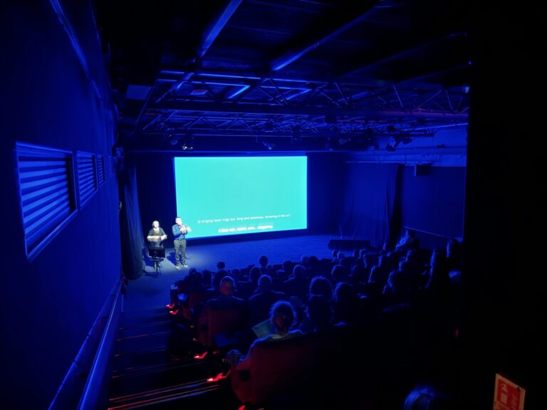 ‘The Blue Description Project’ at the BFI in the UK in March. A group of people are gathered in a cinema. They are seated and are facing a large sized screen. A single shot of saturated blue colour fills the screen and bathes the room and the guests in a blue glow. A caption is seen on the bottom of the screen but is not legible in this image. Two people are standing to the left of the screen as the group faces it.