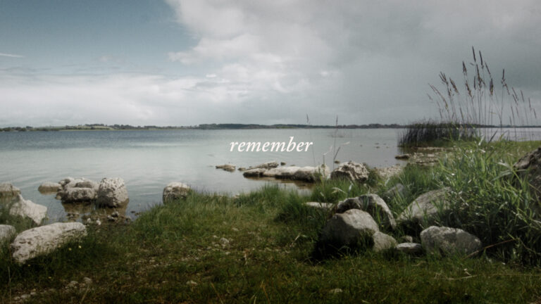 A colour image at the edge of a large mass of water, possibly a lake or river. There are rocks and grass and reeds on either side. The sky is cloudy with a break in the clouds on the left. In the middle of the image is the word remember in white text.