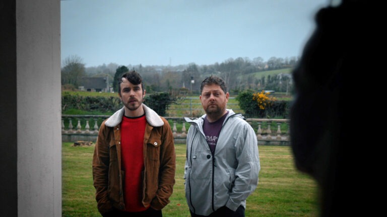 Two men are standing facing the viewer (possibly in a doorway) another person is seen in profile, as a shadow on the right. The man on the left is wearing a red jumper and a rust coloured jacket. The man on the left is wearing a light grey windbreaker and a purple tshirt. They both have their hands in their pockets. In the background there are fields and cow gates, suggesting a rural neighbourhood.