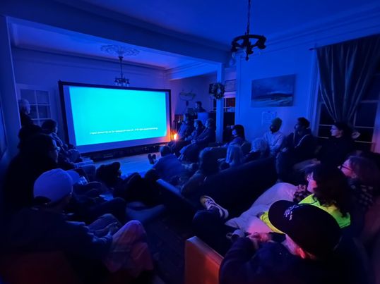 A group of people are gathered in a room. They are mostly seated and are facing a medium sized screen. A single shot of saturated blue colour fills the screen and bathes the room and the guests in a blue glow. A caption is seen on the bottom of the screen but is not legible in this image. 