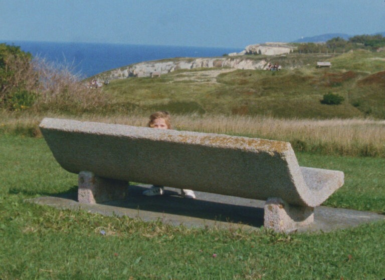 A child is just seen peering over a stone bench which is pictured in a lush green landscape with the sea seen in the distance.