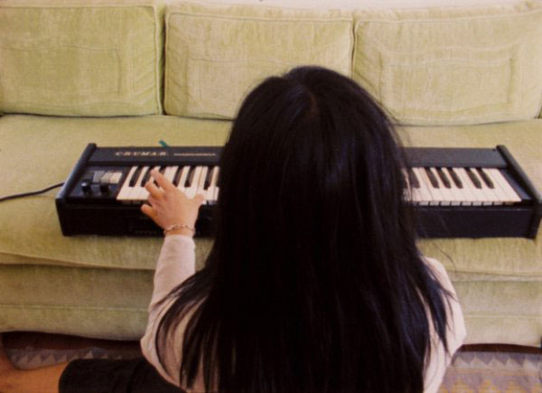 A person with long dark hair is playing a keyboard which has been placed on a cream colored sofa. The person has their back turned to the camera, facing the keyboard and the wall.